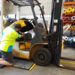 A man is servicing a forklift with an absorbent spill mat under the forklift to catch any leaks