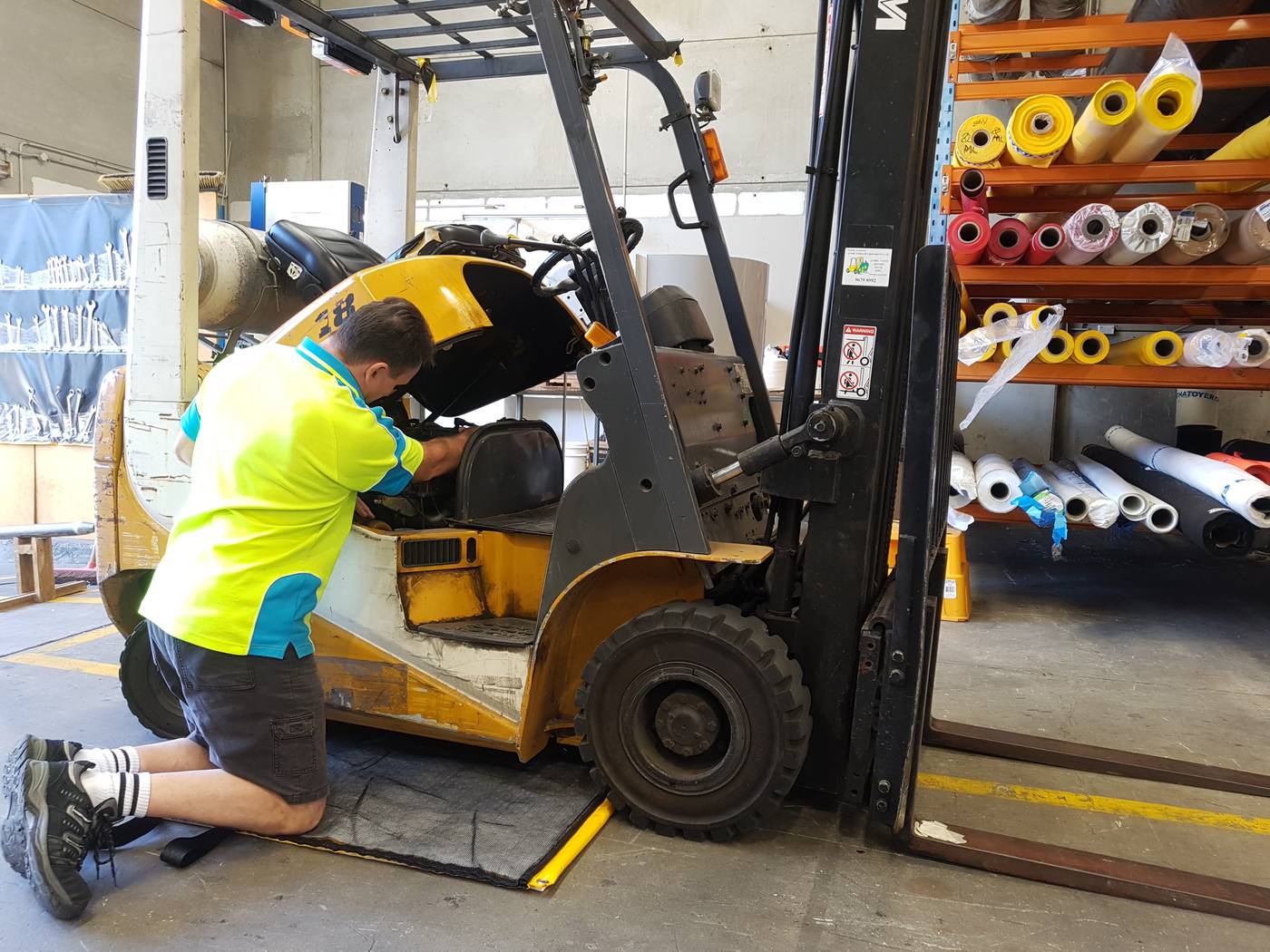 A man is servicing a forklift with an absorbent spill mat under the forklift to catch any leaks
